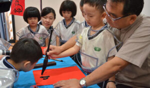 書法老師曾慶仁握著小朋友的手，寫出「春」的象形字，其他孩子也在一旁圍觀。【攝影者：劉曉秀】